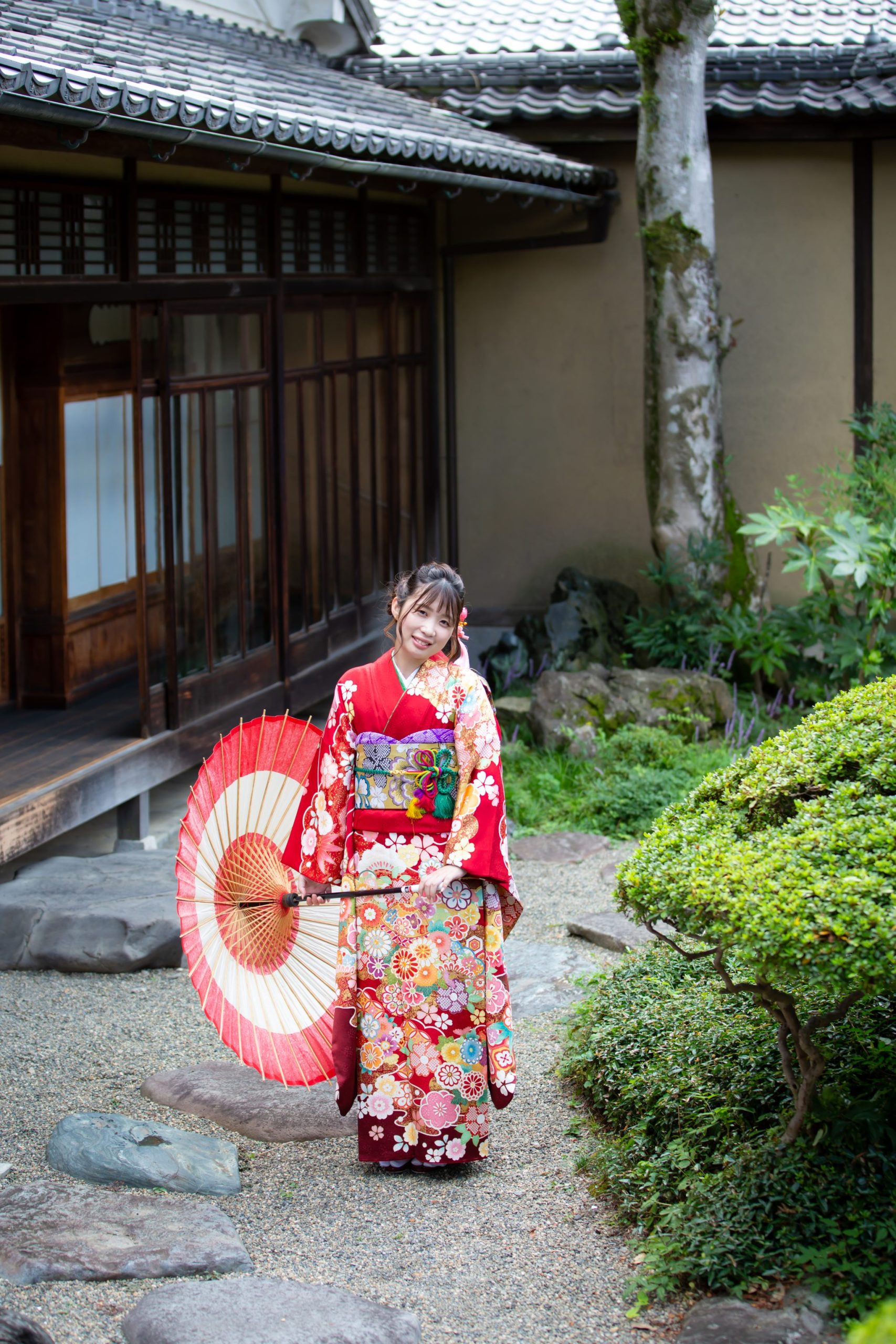 赤い振袖姿で和傘を持ち、日本庭園に立つ女性